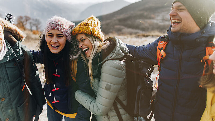 Vier in Winterjacken und Mützen gekleidete Freunde lachen gemeinsam im Freien und halten sich gegenseitig in den Armen. Im Hintergrund sind Berge und ein bewölkter Himmel zu sehen.