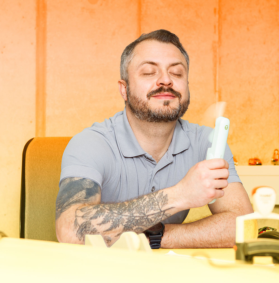 Ein Mann mit Bart und Tattoos sitzt lächelnd an einem Schreibtisch. Er hält sich einen kleinen Handventilator vors Gesicht.