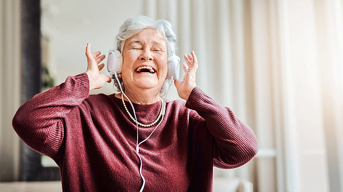 Eine ältere Frau mit grauem Haar, die einen kastanienbraunen Pullover und eine Perlenkette trägt, lächelt fröhlich mit geschlossenen Augen, während sie in einem Haus mit weißen Kopfhörern Musik hört.