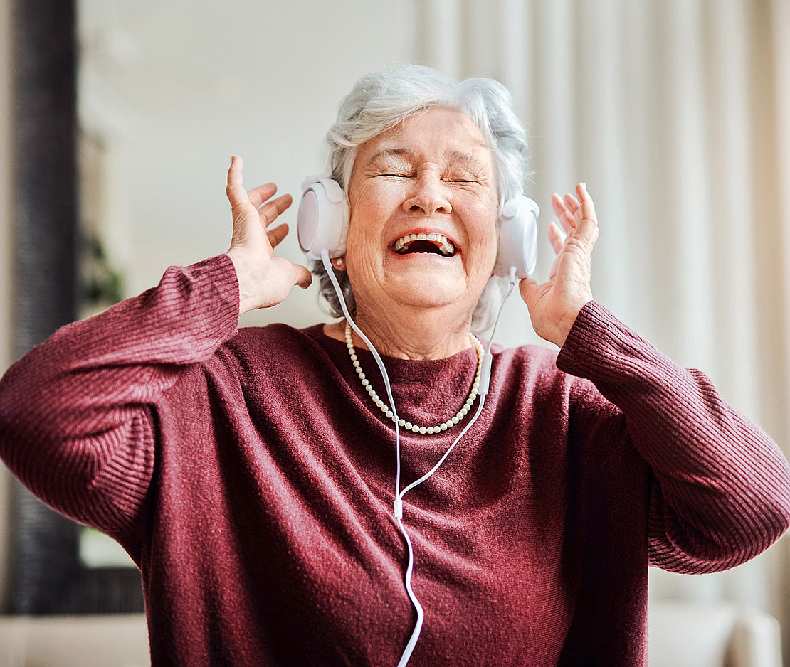 Eine ältere Frau mit grauem Haar, die einen kastanienbraunen Pullover und eine Perlenkette trägt, lächelt fröhlich mit geschlossenen Augen, während sie in einem Haus mit weißen Kopfhörern Musik hört.