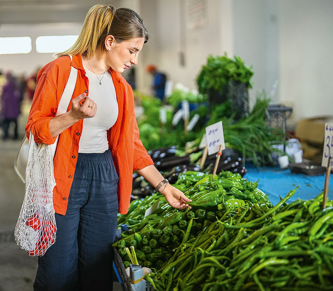 Eine Frau in einem orangefarbenen Hemd begutachtet grüne Paprika an einem Marktstand in einer Halle und hält eine wiederverwendbare Netztasche. Im Hintergrund sind andere Einkäufer und frisches Gemüse zu sehen.