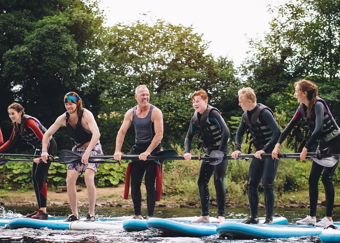 Sechs Personen in Neoprenanzügen und Schwimmwesten stehen auf Paddelbrettern, lächeln und halten Paddel, auf einem Fluss mit Bäumen im Hintergrund.