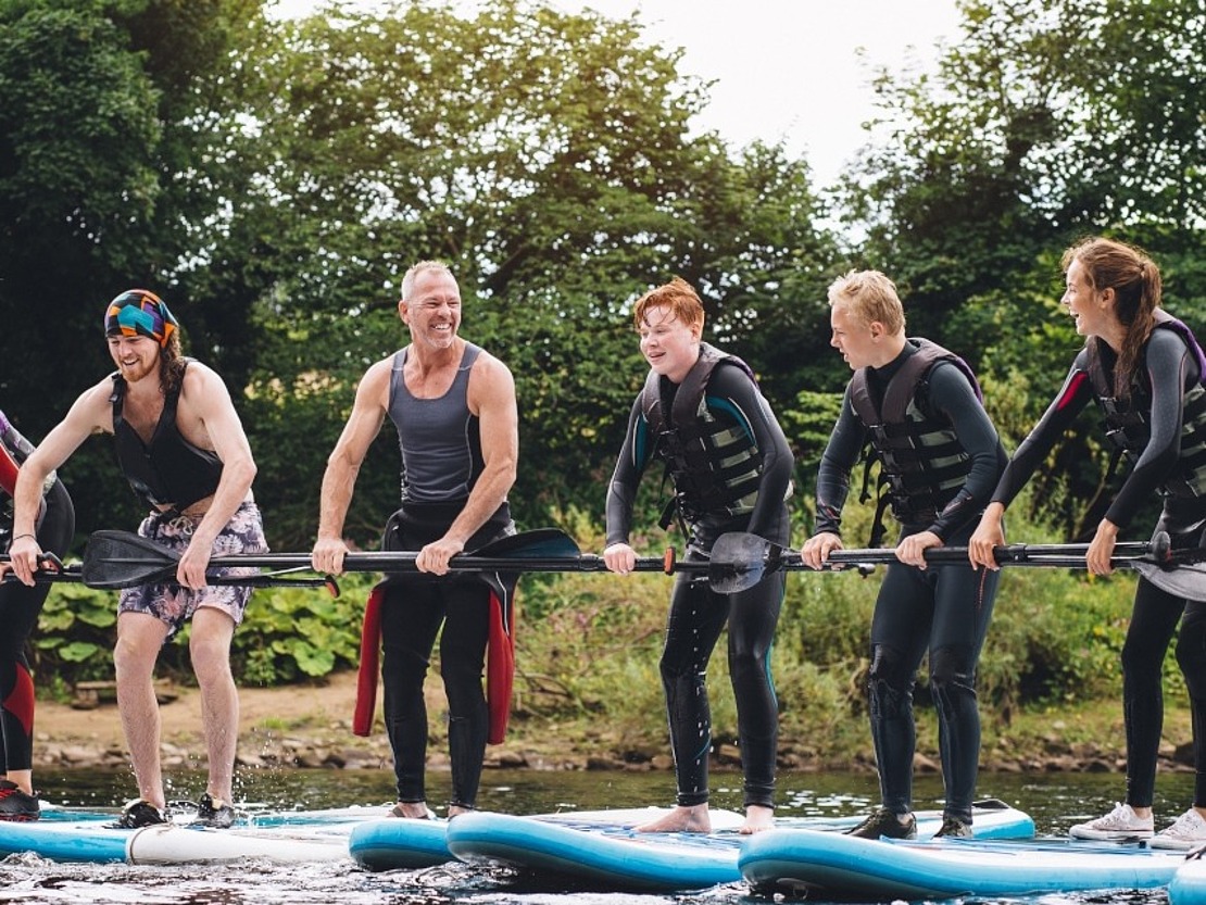 Sechs Personen in Neoprenanzügen und Schwimmwesten stehen auf Paddelbrettern, lächeln und halten Paddel, auf einem Fluss mit Bäumen im Hintergrund.