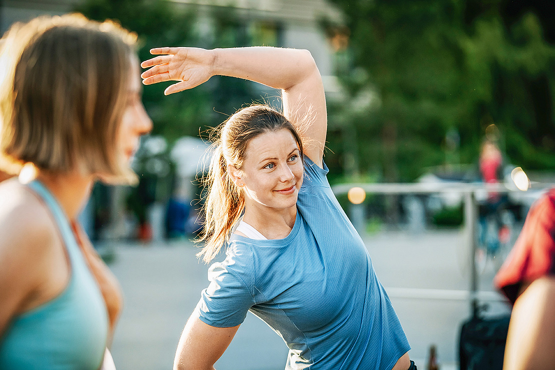 Eine Frau in einem blauen Hemd streckt ihren Arm über den Kopf, während sie im Freien trainiert, lächelt und schaut zur Seite. Eine andere Person in Trainingskleidung ist unscharf im Vordergrund zu erkennen. Bäume und Strukturen im Freien sind im Hintergrund zu sehen.