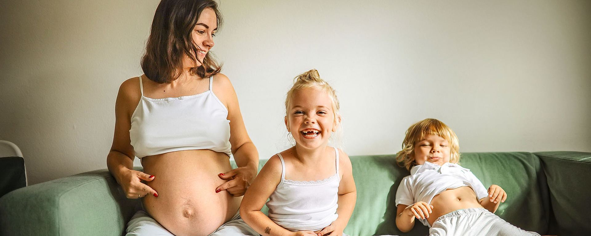 Eine schwangere Frau sitzt mit zwei kleinen Kindern auf einem grünen Sofa. Alle drei heben ihre Hemden hoch, um ihre Bäuche zu zeigen, und lächeln verspielt. Das sorgt für einen fröhlichen und entspannten Familienmoment.