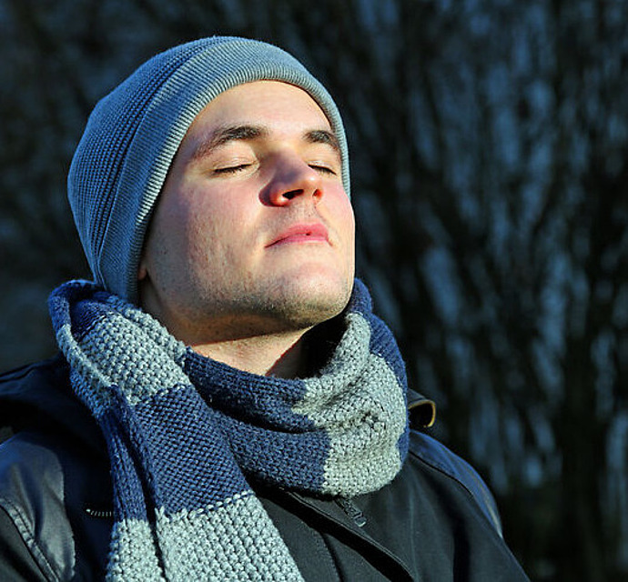 Ein junger Mann mit grauer Strickmütze und Schal steht im Freien, die Augen geschlossen, dem Sonnenlicht zugewandt und wirkt ruhig und zufrieden. Die dunklen Bäume im Hintergrund sind unscharf.