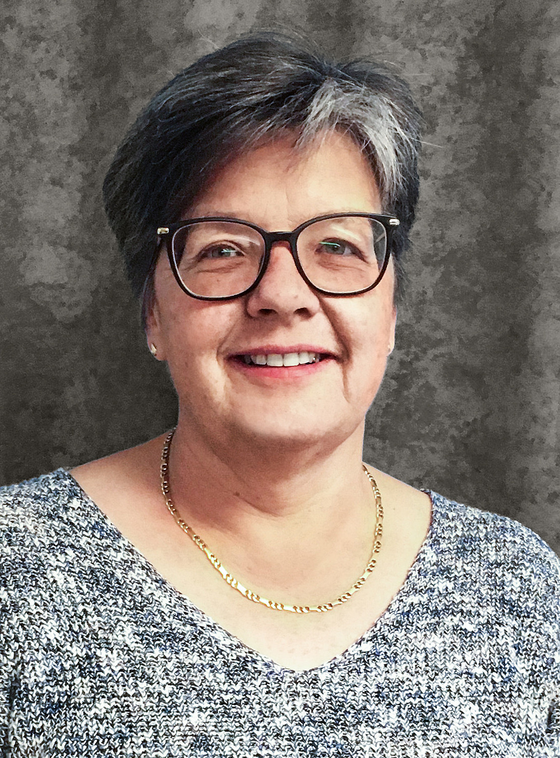 Eine lächelnde Frau mit kurzen grauen Haaren und Brille, die eine goldene Halskette und einen grau gemusterten Pullover trägt, posiert vor einem grau-melierten Hintergrund.