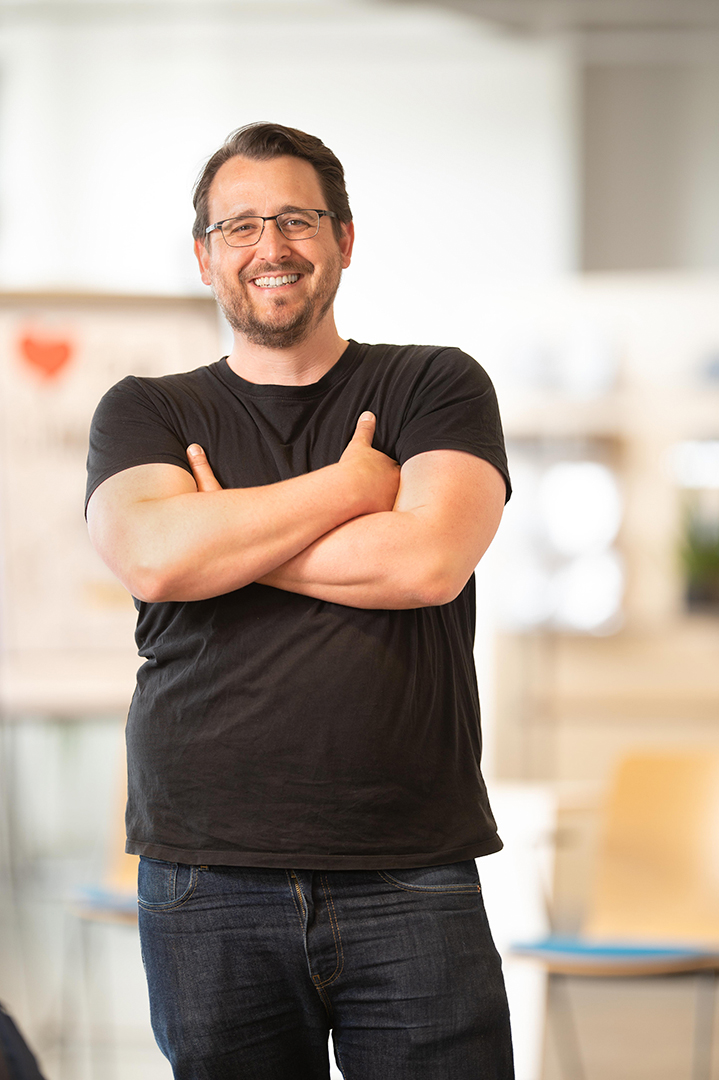 Ein lächelnder Mann mit Brille, braunem Haar und Bart steht mit verschränkten Armen in einem Raum und trägt ein schwarzes T-Shirt und Jeans. Der Hintergrund ist leicht unscharf und zeigt Stühle und eine helle, moderne Umgebung.