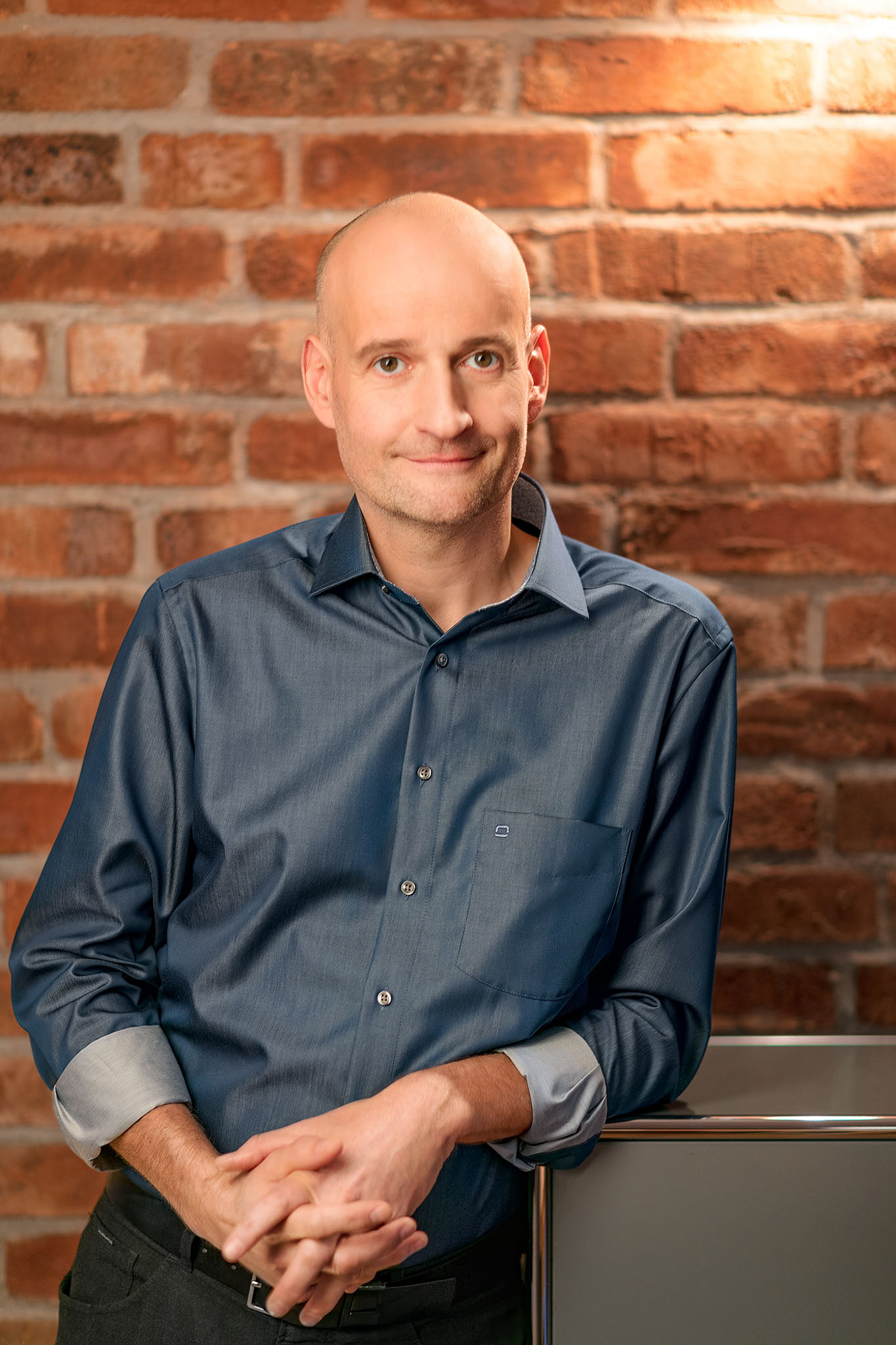 Ein Mann mit rasiertem Kopf und blauem Hemd steht vor einer Backsteinmauer. Er lächelt leicht und hat die Hände vor der Brust gefaltet. Warmes Licht hebt die Szene hervor.