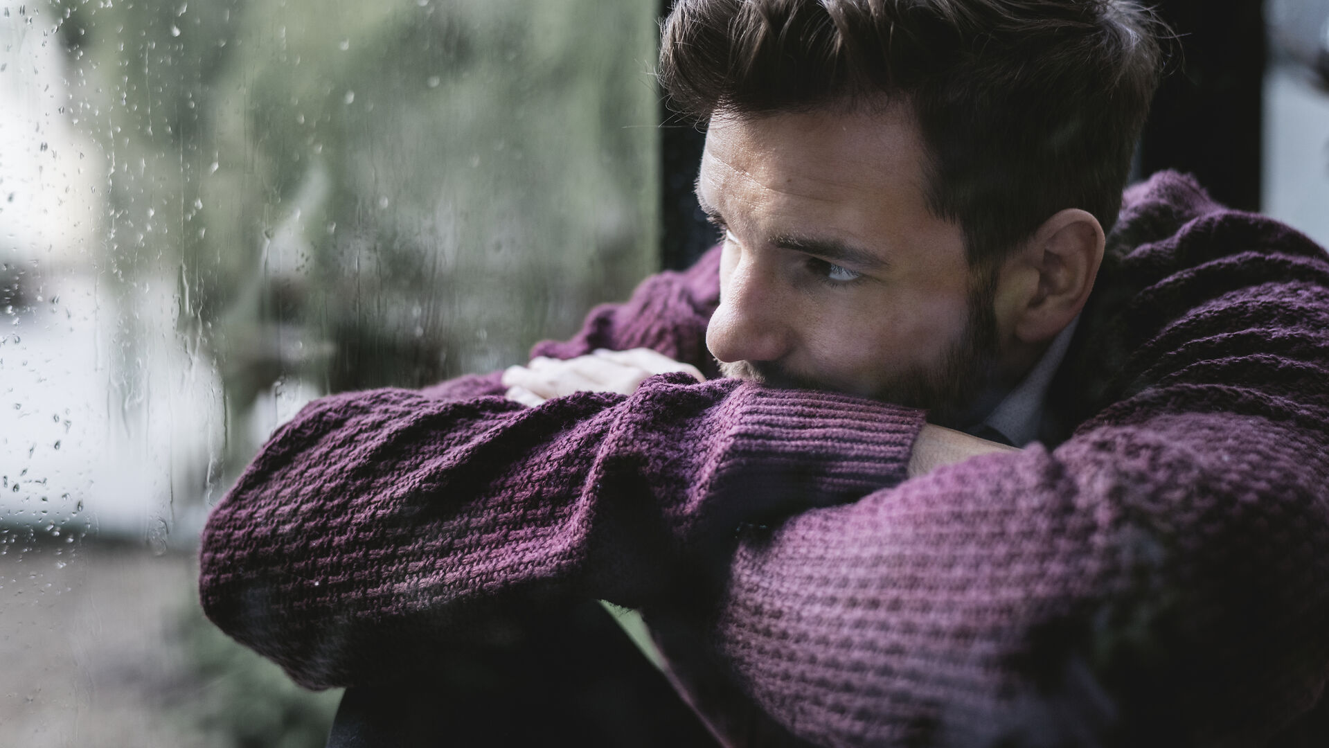 Ein Mann in einem kastanienbraunen Pullover sitzt in einem Haus, stützt die Arme auf die Knie und blickt nachdenklich aus einem regengesprenkelten Fenster. Die Stimmung ist kontemplativ und düster.